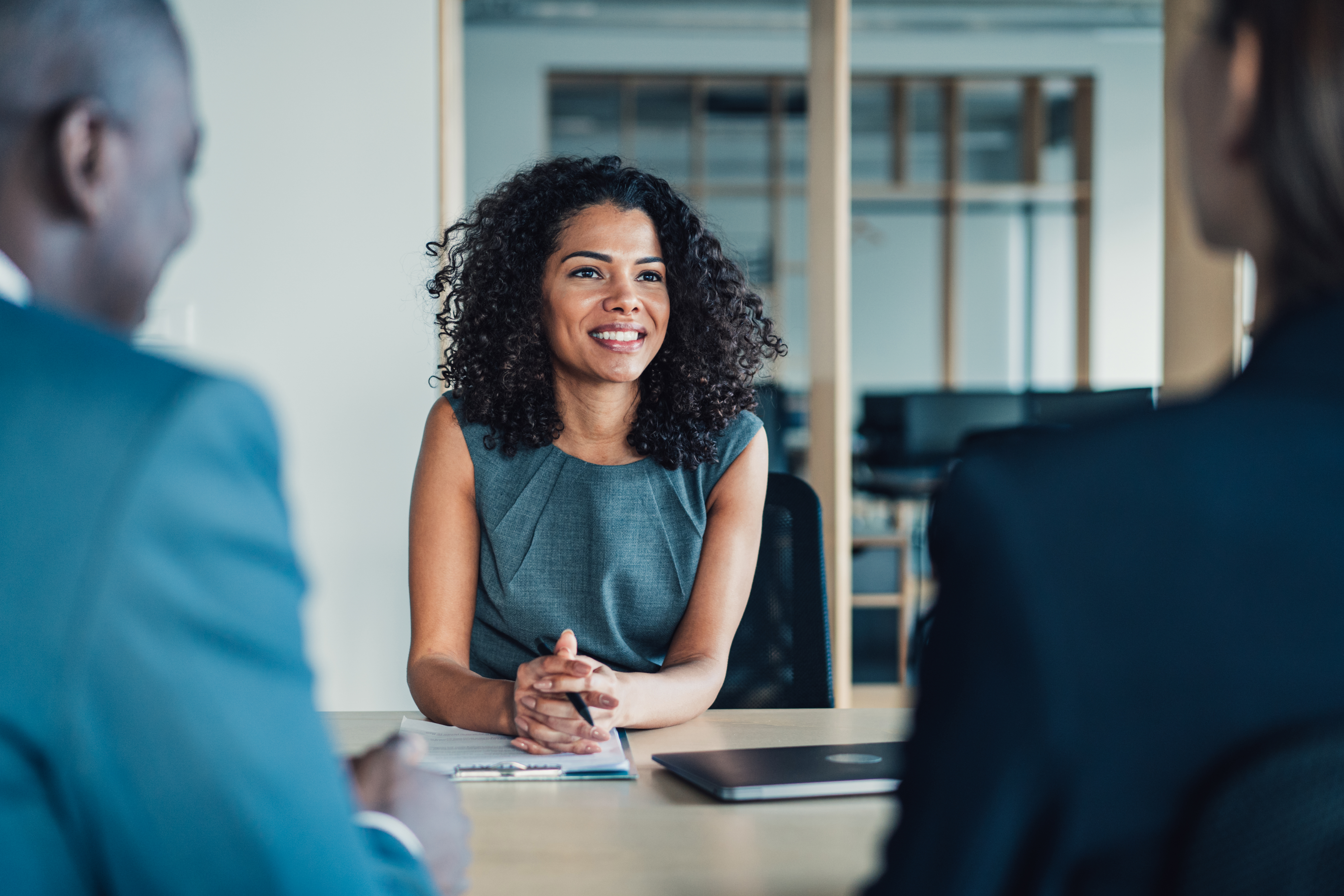 A smiling woman is sitting at a table in a meeting room, opposite two colleagues in suits