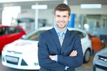 A smiling man in a navy suit, light blue shirt, and striped tie is standing in front of vehicles with his arms crossed