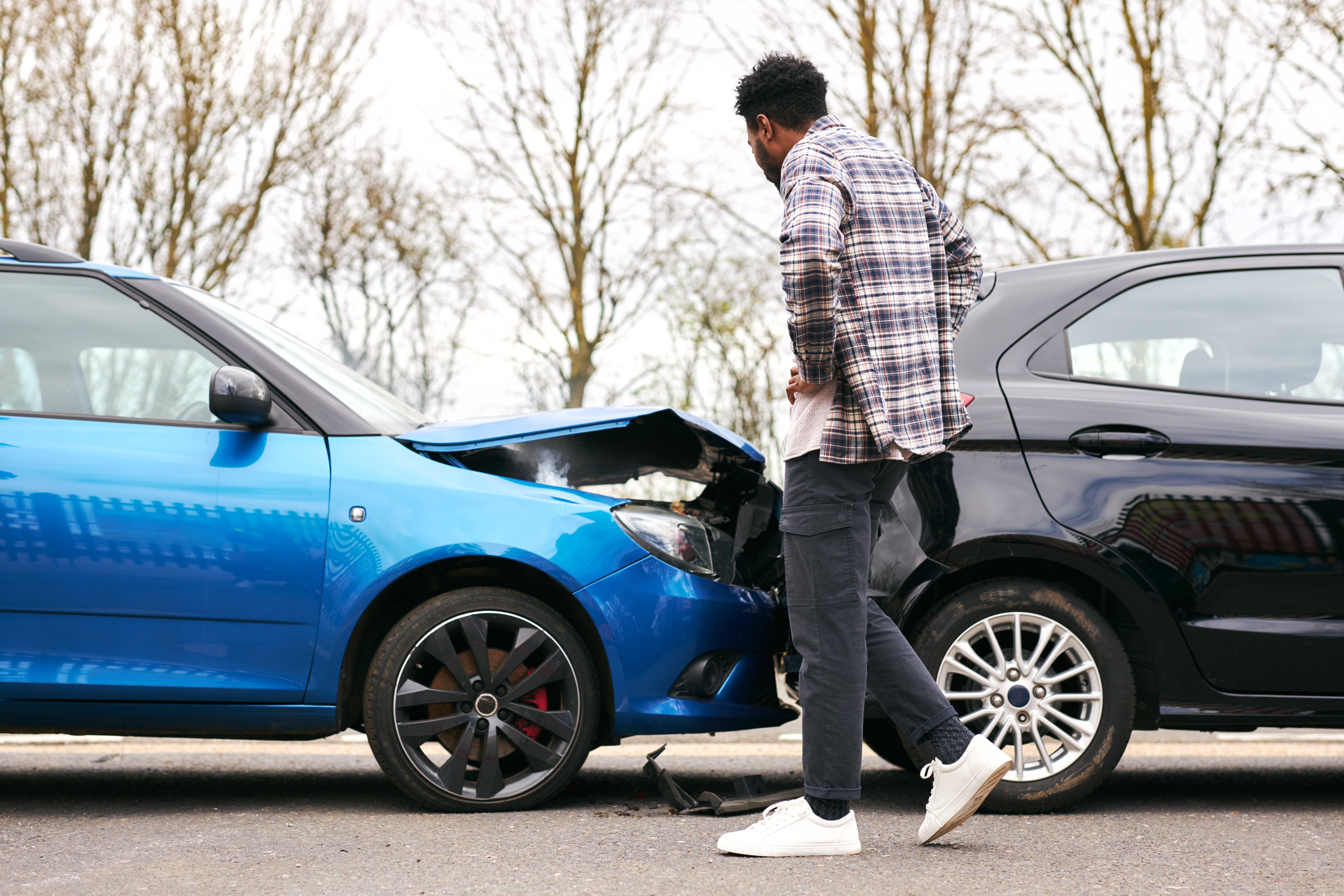 A man stands with hands on hips in front of a blue car that has gone into the back of a black car