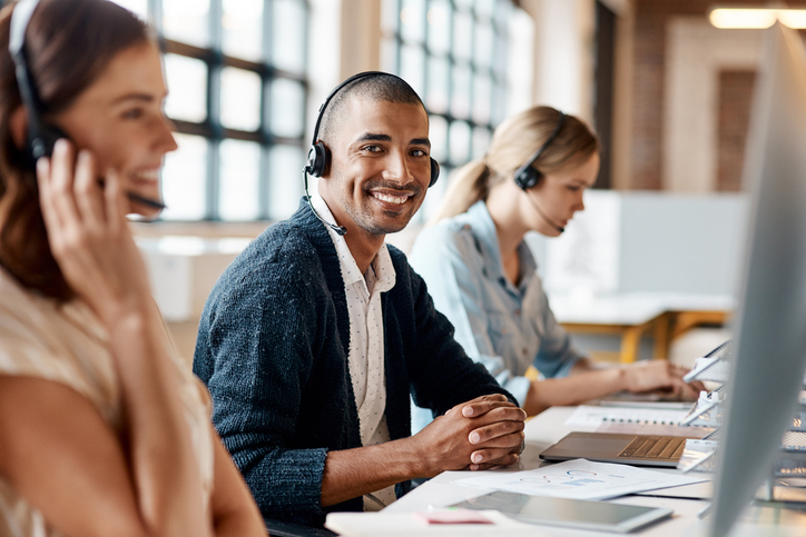 A man is wearing a headset at a desk, in front of his laptop, smiling at the camera. Two women in headsets are working next to him
