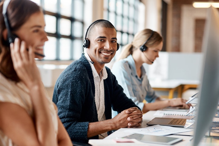 A man is wearing a headset at a desk, in front of his laptop, smiling at the camera. Two women in headsets are working next to him