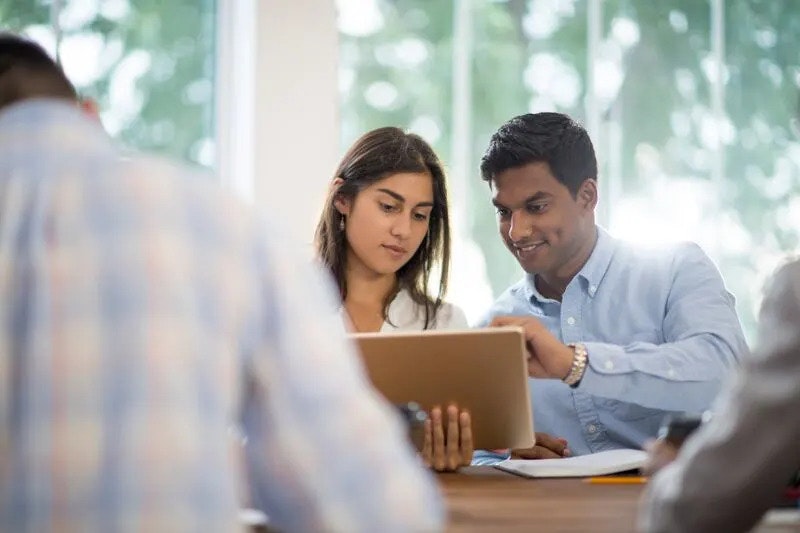 A woman and a smiling man sitting at a table and looking at a screen which the woman is holding and showing to the man