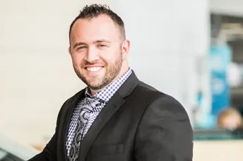 A man in a dark suit with a chequered purple shirt and tie is smiling at the camera