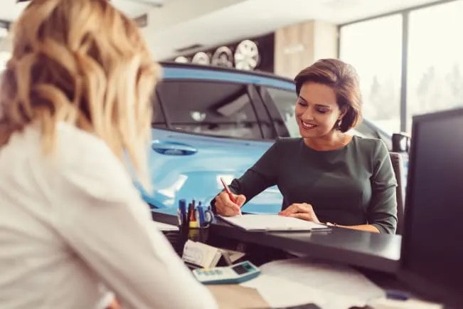 A female worker at her desk in a dealership. A woman is opposite her, signing paperwork