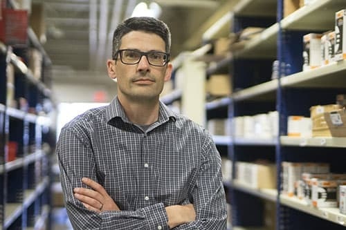 A man in a chequered black and white shirt is looking at the camera, arms crossed, behind him are shelves with parts